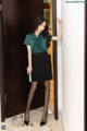 A woman in a green shirt and black skirt leaning against a door.