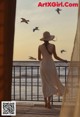 A woman in a white dress and hat standing on a balcony looking out at the ocean.