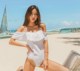 A woman in a white one piece swimsuit sitting on a beach.