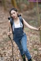 A woman in overalls and a backpack standing in the woods.