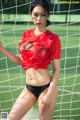 A woman in a red shirt and black panties posing in front of a soccer goal.