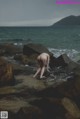 A naked woman standing on a rocky beach next to the ocean.